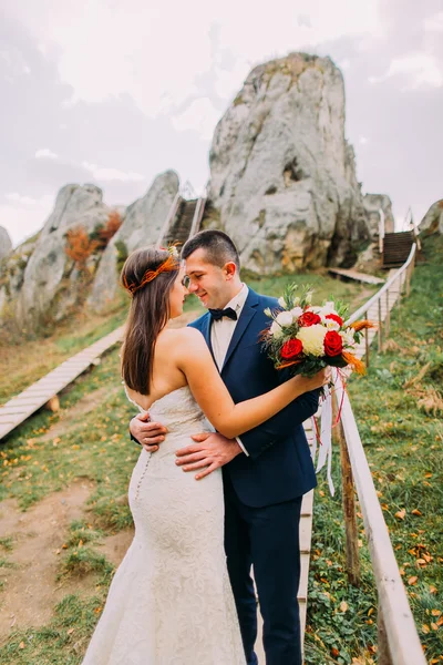 Handsome groom in stylish blue suit gently embracing his white dressed bride holding bouquet of roses on majestic mountain landscape with big rocks as backround