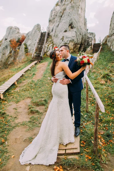 Handsome groom in stylish blue suit kissing his white dressed bride holding bouquet of roses on majestic mountain landscape with big rocks as backround