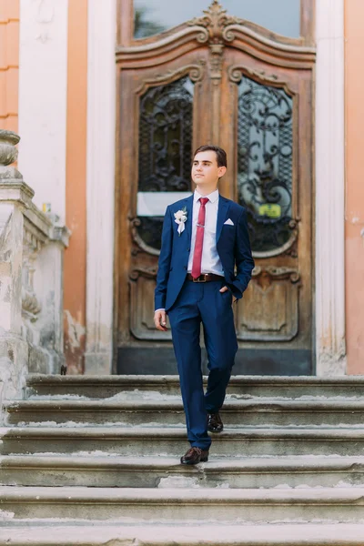 Young and confident newlywed groom posing in blue suit on old stairs at vintage building entrance