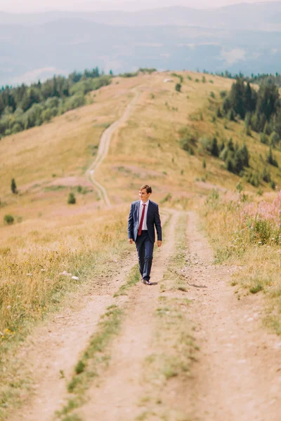 Young man in stylish suit walking on trail by summer field with hills at background