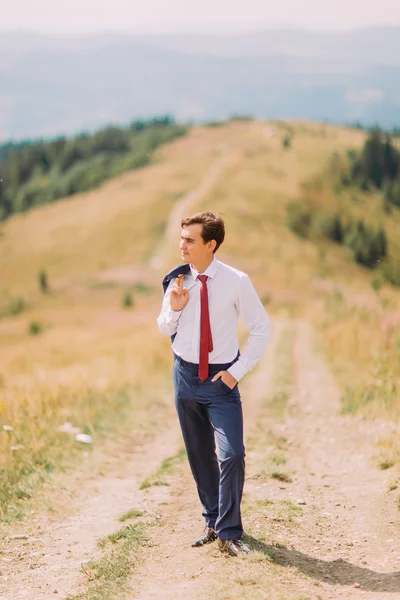 Young man in stylish suit walking on trail by summer field with his blazer over shoulder. Forest hills at background