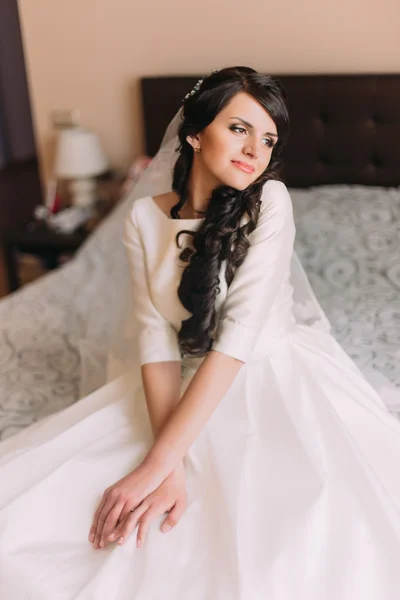 Young fashionable excited bride sitting on bed in wedding dress and dreaming of her new married life