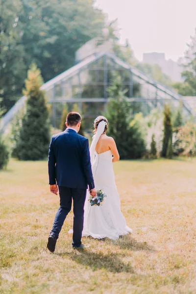 Beautiful bride with white wedding dress and groom in stylish blue suit walking outdoor on lawn. Greenhouse at background