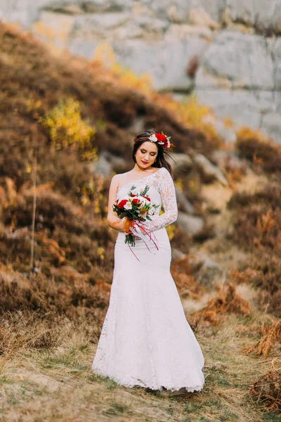 Innocent romantic beautiful bride in luxurious wedding dress. Young girl with bouquet of red white roses and head wreath. Rocky landscape as background