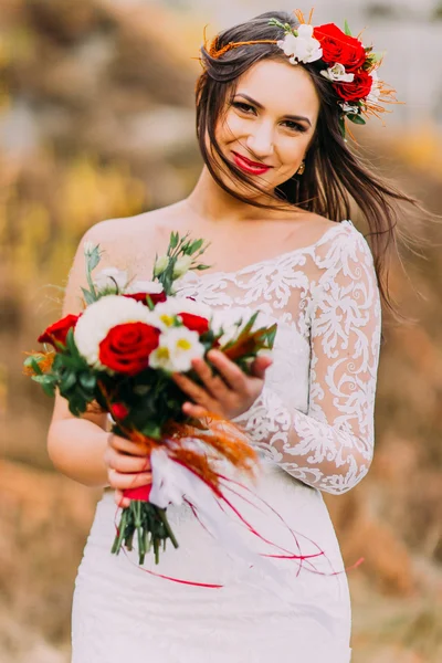 Innocent romantic beautiful bride in stylish wedding dress. Young girl with bouquet of red white roses and head wreath. Natural landscape as background