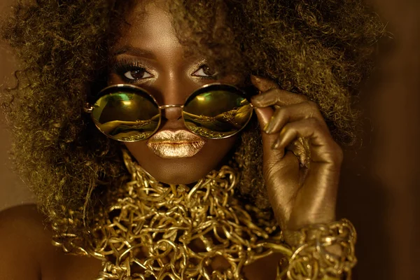 Close-up of magic golden african american female model in massive sunglasses with bright glitter makeup, glossy hairstyle and big red lips posing on the studio background