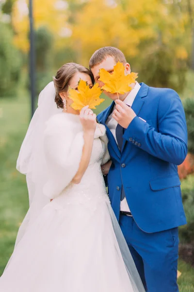 Newly married couple posing outdoors. Young people hiding their faces behind autumn leaves