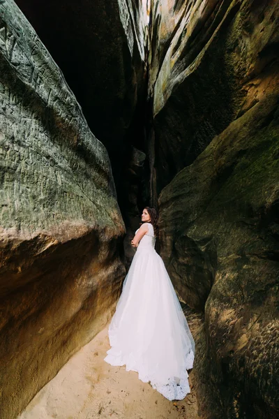 Beautiful and innocent bride in long-tailed white dress posing at darkened rock cleft