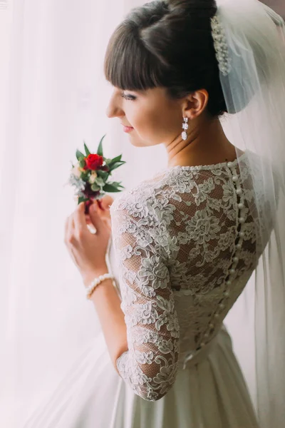 Thoughtful young bride posing with cute floral boutonniere near the window before wedding ceremony