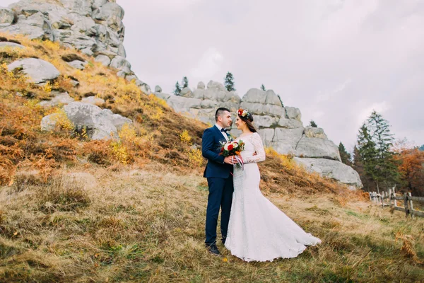 Handsome groom in stylish blue suit embracing white dressed bride holding bouquet of roses on majestic landscape with rocks and fence as backround