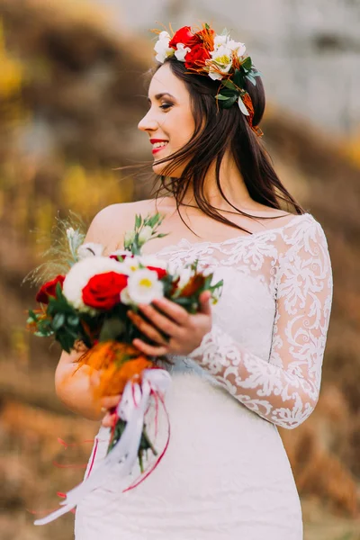 Innocent romantic beautiful bride in stylish wedding dress. Young smiling girl with bouquet of red white roses and head wreath. Natural landscape as background