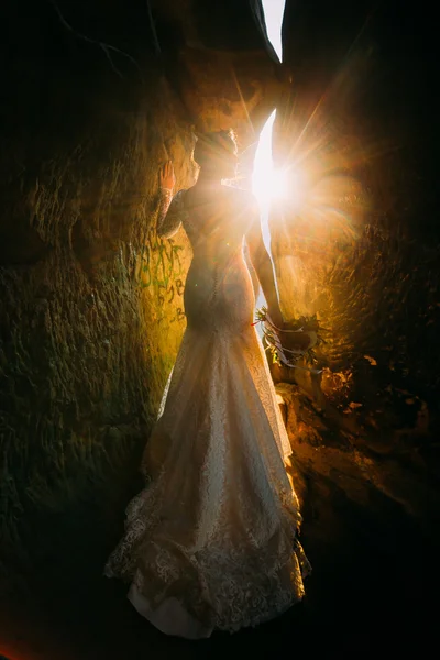 Silhouette of beautiful young woman wearing elegant white dress standing between two rocks with yellow sunset rays beaming through the cleft