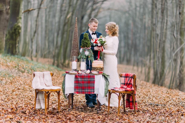 Happy young wedding couple near the decorated table for Christmas holidays in autumn forest.