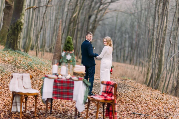 Happy young wedding couple walking and holding hands near the decorated table for Christmas holidays in autumn forest.