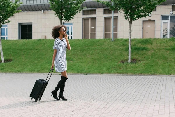 Full body portrait of a traveling african-american woman walking with suitcase and mobile phone