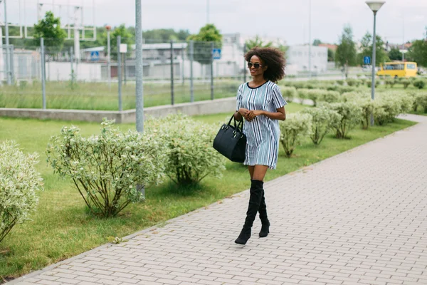 Full length portrait of an smiling young african american woman in short dress and jackboots walking with a purse looking ahead