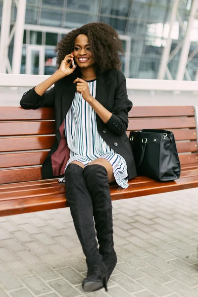 Happy African American woman in dress and jackboots using cellphone sitting on bench while waiting for flight