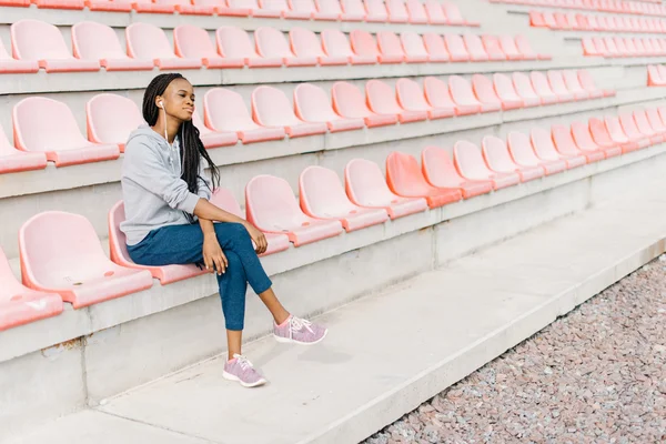 Slim relaxing african american female in sportswear sitting on plastic red seat at the stadium and listening to music