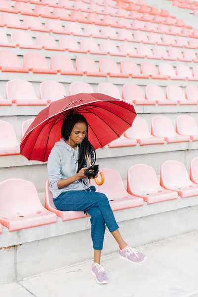 The beautiful young african american woman with phone, in earphones holding red umbrella. Sport at stadium