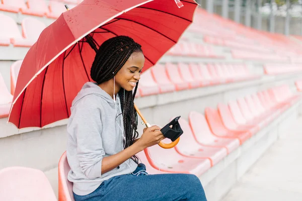 Young african american girl in sport suit with red umbrella sitting on the stadium. She listening to music