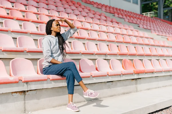 Full length of pretty amusing young woman sitting on modern stadium benches and looking far away