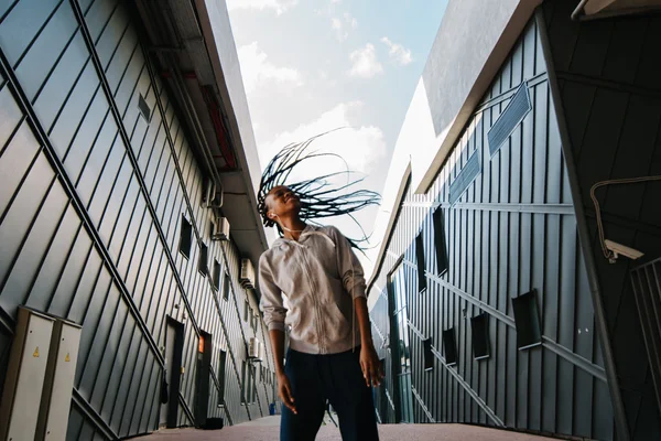 Young african American woman listening to music and swinging her braided hair, modern buildings at background