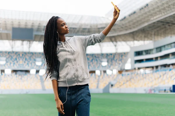 Young beautiful african american sportswoman making selfie photo on mobile phone after training outdoors and smiling. Fit woman is at large nice modern stadium
