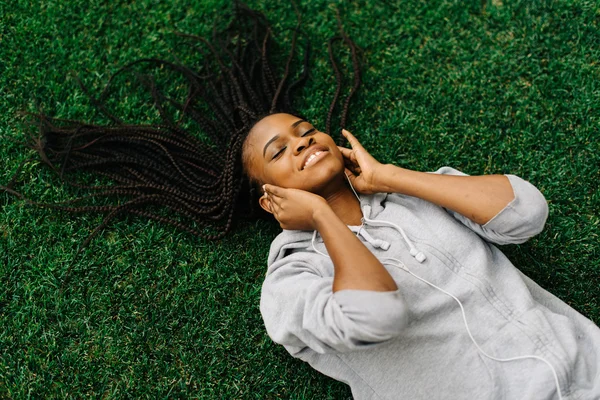 Close up portrait of a beautiful and dreamy African American woman laying down on healthy green grass, relaxing with her eyes closed, smiling outdoors