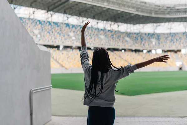 Back of african american woman standing among empty stadium celebrating, her arms raised punching the air with hands