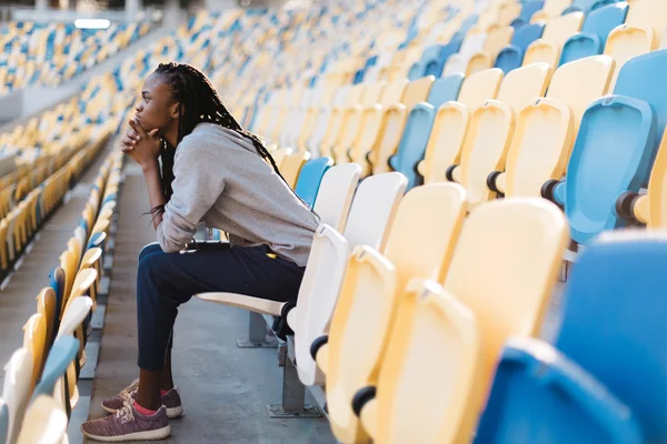 Side view of sad african american young woman sitting at stadium with her chin resting on hands and a glum expression