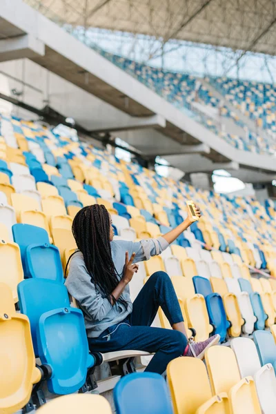 Portrait of happy young woman gesturing victory or peace hand sign taking a selfie with her cellphone at stadium