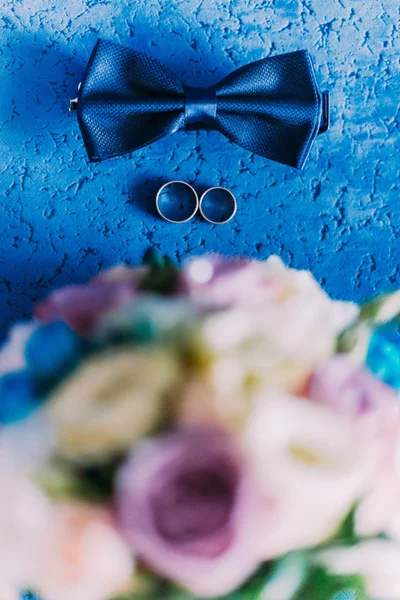 Still life with bow tie, wedding bouquet and two golden wedding rings on blue background