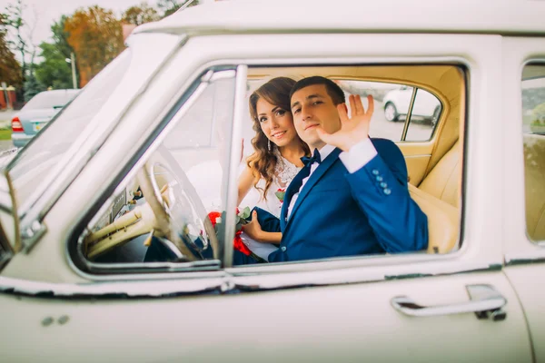 Happy newlywed couple sitting in vintage car. Groom is waving his hand to the attendants