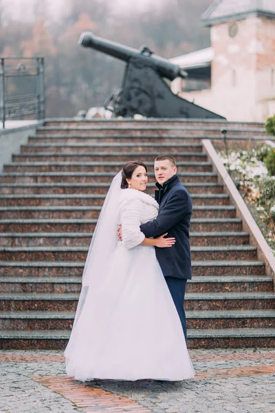 Beautiful bride in long white dress and elegant groom holding each other on stairs outdoors. Old cannon at background