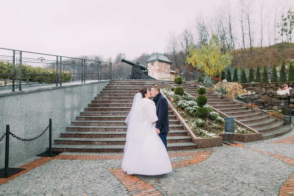 Beautiful bride in long white dress share kiss with elegant groom on stairs outdoors. Old cannon at background