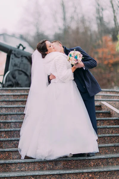 Beautiful bride in long white dress kissing with elegant groom on stairs outdoors. Old cannon at background