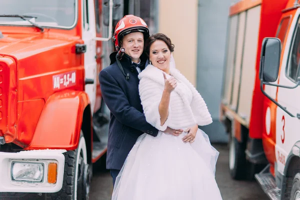 Happy wedding couple posing near big red fire truck. Groom in firemans helmet