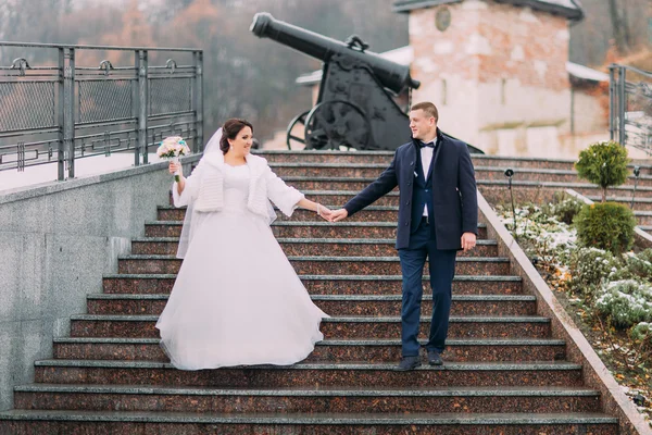 Beautiful bride in long white dress walking with elegant groom on stairs outdoors. Old cannon at background