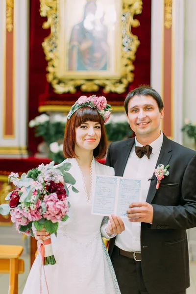 Gorgeous emotional red-head bride and handsome young groom holding their certificate in church
