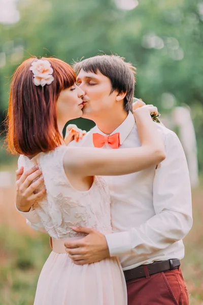 Groom kisses bride tenderly holding her waist while they stand in the park