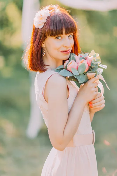 Young red-haired bride in simple lace dress with bouquet of pink flowers She smiles softly. On her head - wreath