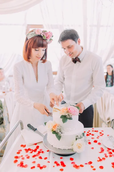 Charming bride and handsome groom are cutting a wedding cake. Couple in the restaurant with white pie