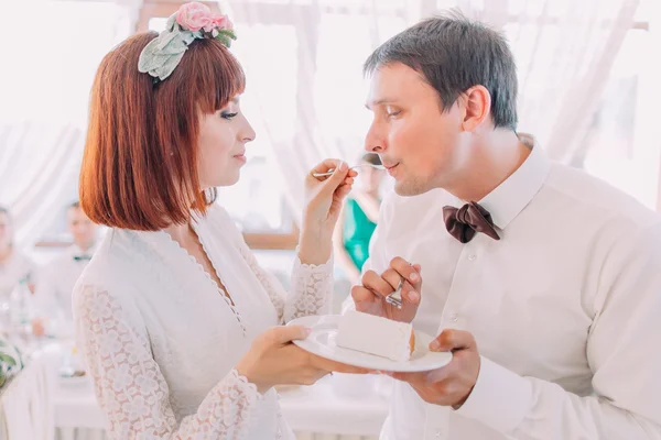 Beautiful young bride feeding wedding cake to groom in light restaurant interior
