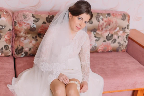 Beautiful bride sitting on sofa in dressing room dresses bridal garter, preparing herself for wedding ceremony