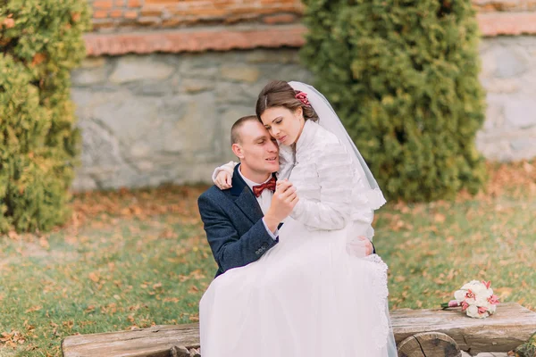 Happy newlywed couple in autumn park. Elegant bride sitting on knees of her handsome new husband