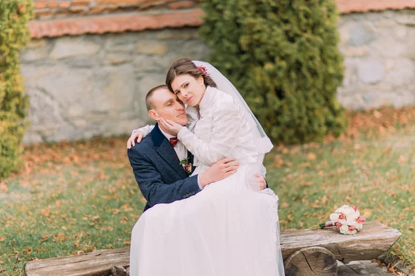 Happy just married couple in autumn park. Elegant bride sitting on knees of her handsome new husband