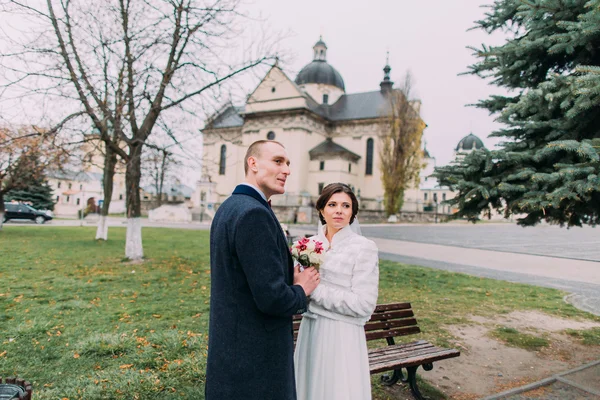 Panoramic view of beaytiful newlywed couple holding hands and looking afar in park near old baroque church