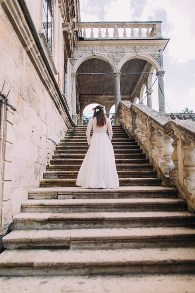 Charming bride in long white wedding gown and floral wreath going up by antique palace stone stairs