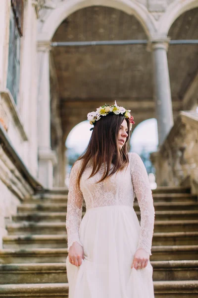 Beautiful young bride in long white wedding gown and floral wreath posing on antique palace stone stairs