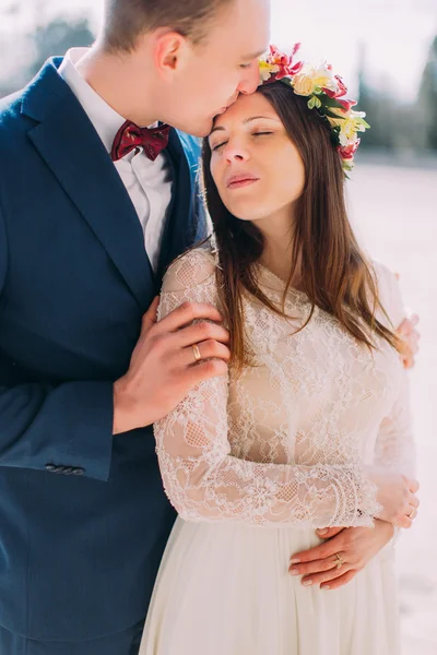 Outdoor portrait of happy sensual wedding couple embracing. Handsome groom holding his pretty new wife and kisses her in forehead
